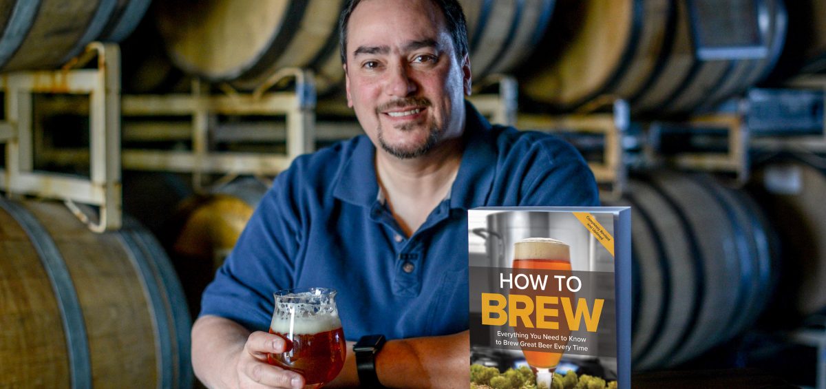 John Palmer, author of How To Brew, at a table in a barrel room with his book and a glass of beer