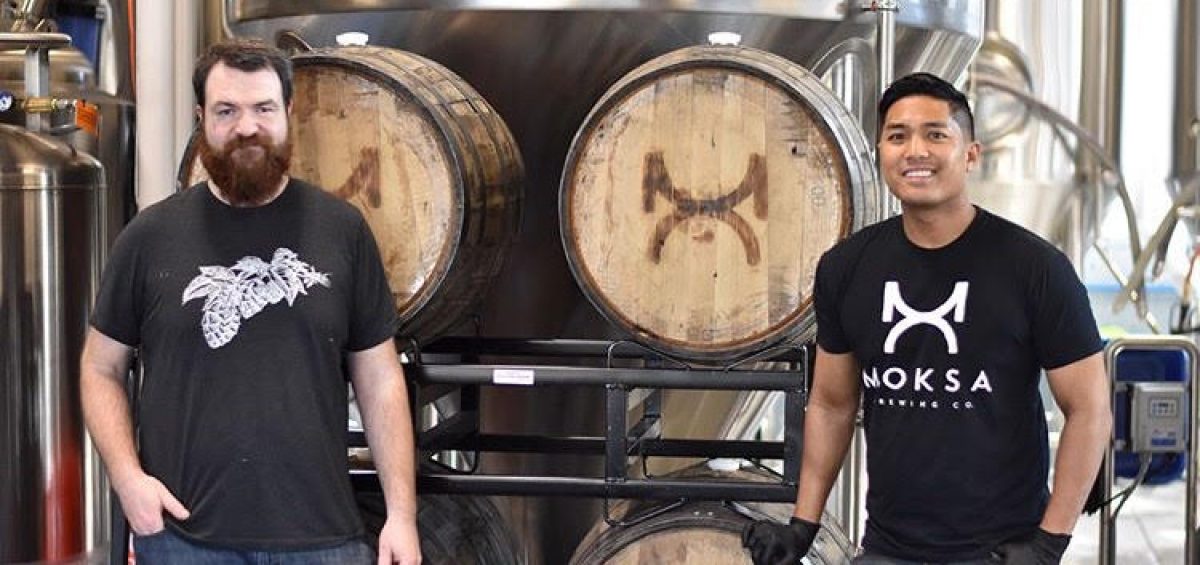 Brewers Derek Gallanosa and Cory Meyer in front of beer barrels at Moksa Brewing Company